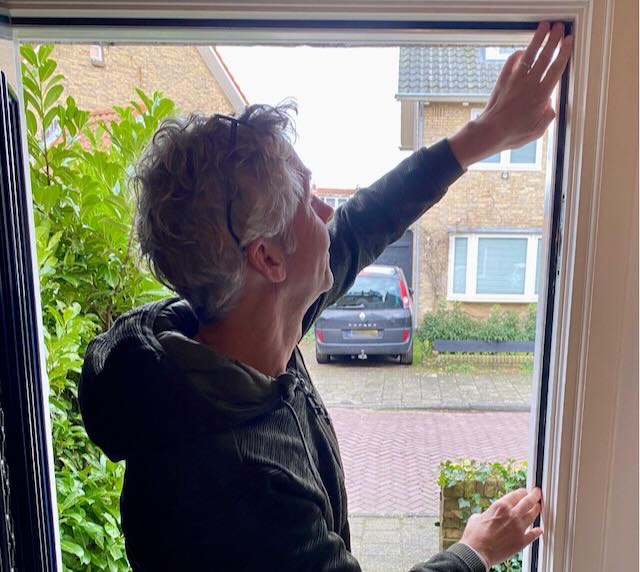 Een bewoner van het Ramplaankwartier drukt tochtstrip aan langs de binnenzijde van een openstaand kozijn, met uitzicht op de straat.