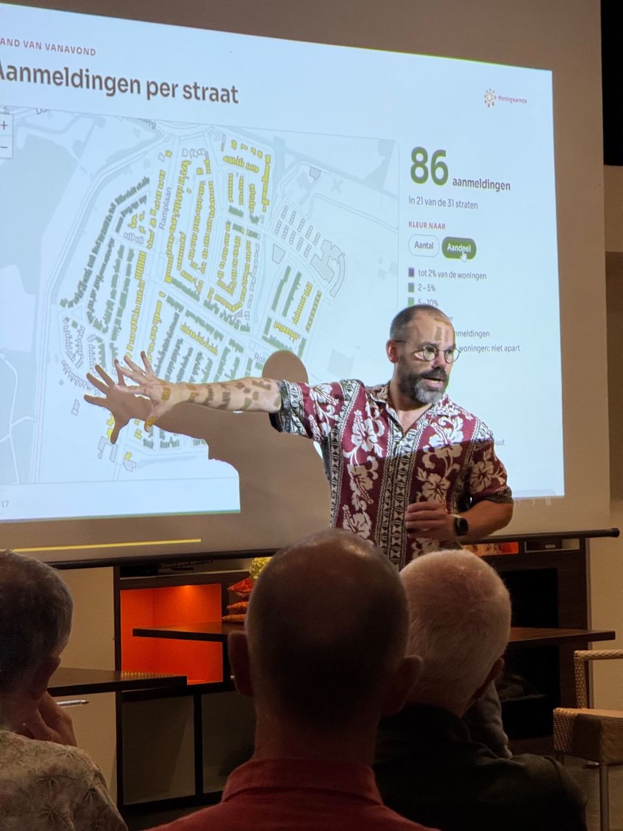 Een teamlid van Woningwarmte wijst tijdens een presentatie naar een kaart van de wijk op het scherm, met de kop Aanmeldingen per straat en het getal 86 aanmeldingen in 21 van de 31 straten.