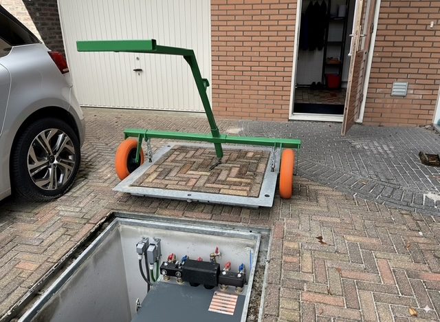Het deksel van een warmtepompkelder hangt opgetild in een hijsframe; in het deksel ligt dezelfde klinkerbestrating als in de oprit, zodat er na afloop niets van te zien is. Daaronder staat de warmtepomp in de kelder.