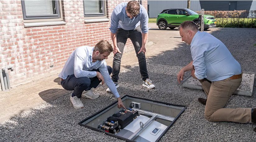 Een geopende warmtepompkelder in een oprit: in de kelderbak onder straatniveau staat de warmtepomp compleet gemonteerd, drie mannen bekijken de installatie.