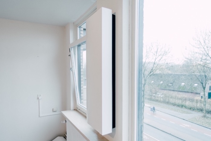 Een smalle, staande witte ventilatie-unit tegen de muur naast een raam, met één doorvoer naar buiten en zonder kanalen door de woning.