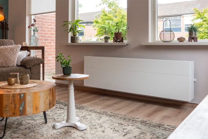 Een brede, lage witte ventilatie-unit onder het raam van een woonkamer, op de plek waar anders een radiator zou hangen.